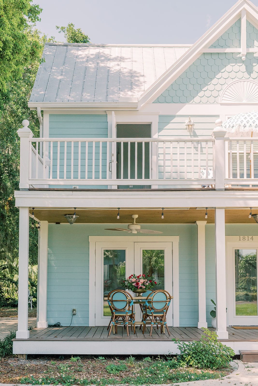 A charming entryway features a covered porch with a small round table surrounded by four chairs. A vibrant floral arrangement sits in the center, while ceiling fans and lights enhance the outdoor space. The light blue exterior contrasts with the greenery surrounding the house.