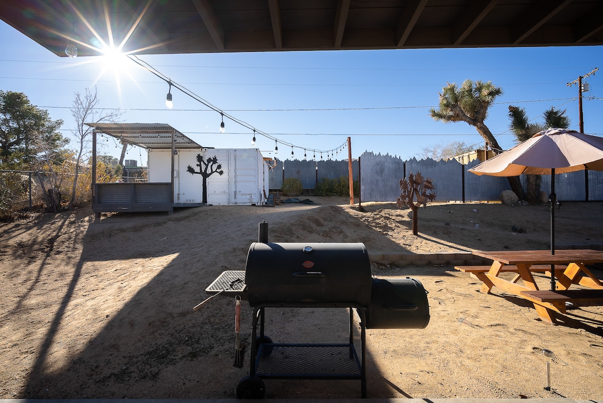 A BBQ grill is positioned in the foreground, with a picnic table nearby. The sunlight creates a bright atmosphere, illuminating the outdoor space, including a shaded area with an umbrella and a distant shipping container. A unique Joshua tree is visible, contributing to the desert landscape.