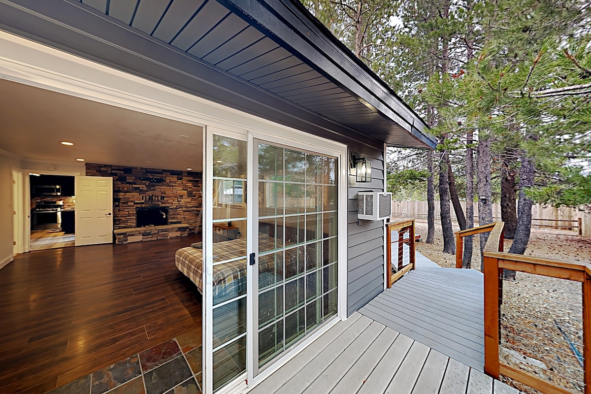 A sliding glass door opens to a spacious living area featuring a stone fireplace. The outdoor deck is surrounded by trees, creating a natural setting. Warm wood tones and a cool grey exterior enhance the inviting atmosphere of this ranch-style home.