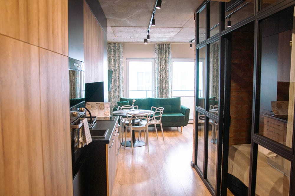 A modern apartment interior features a kitchenette with wood paneling and glossy surfaces. A round dining table with four chairs is positioned beside a plush green sofa. Large windows are dressed with patterned curtains, creating a bright and inviting atmosphere.