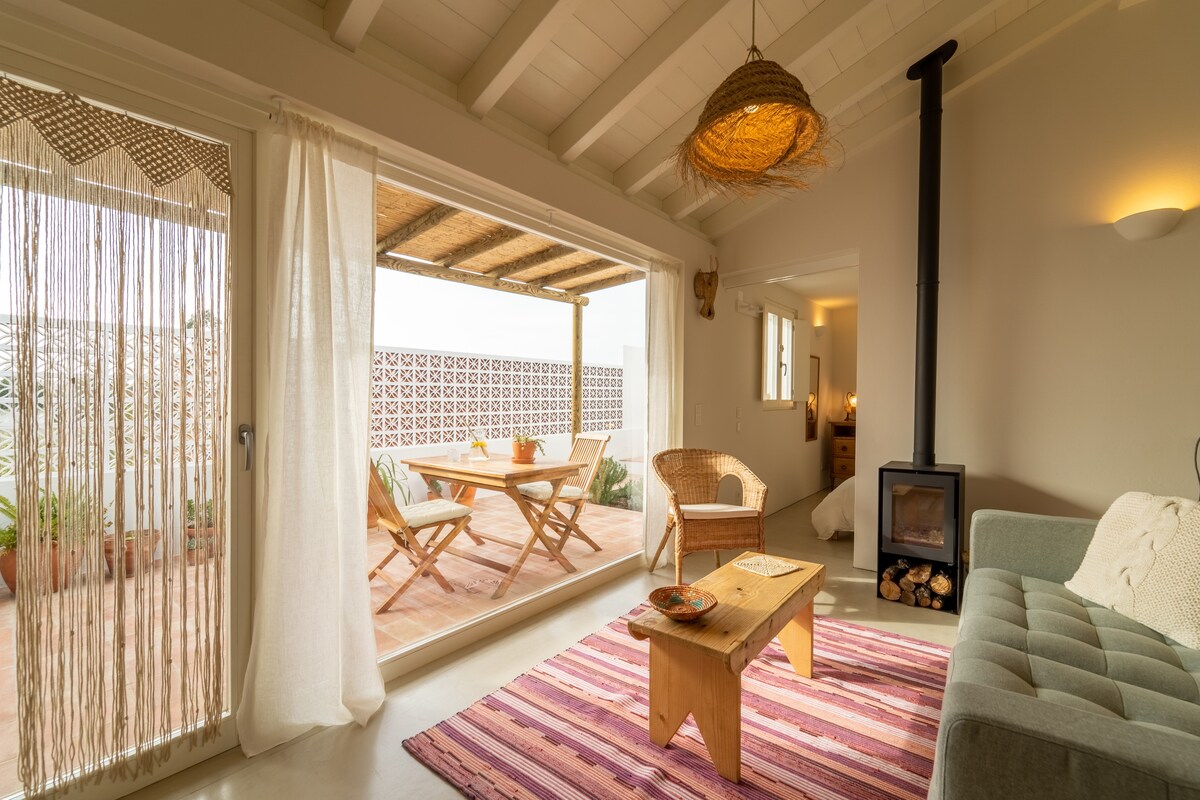 A cozy living area features a wood-burning stove and light-colored furniture. Natural light floods the space through glass doors, leading to an inviting terrace equipped with wooden dining furniture. A woven wall hanging and a textured area rug add warmth to the minimalist decor.