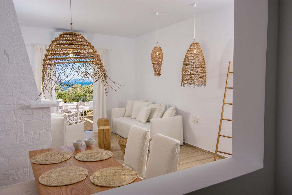 A bright living area features a light-colored sofa and woven pendant lights. Natural textures are introduced through decorative elements and a wooden dining table set for four. A view of the sea is visible through an open door, enhancing the space's tranquility.
