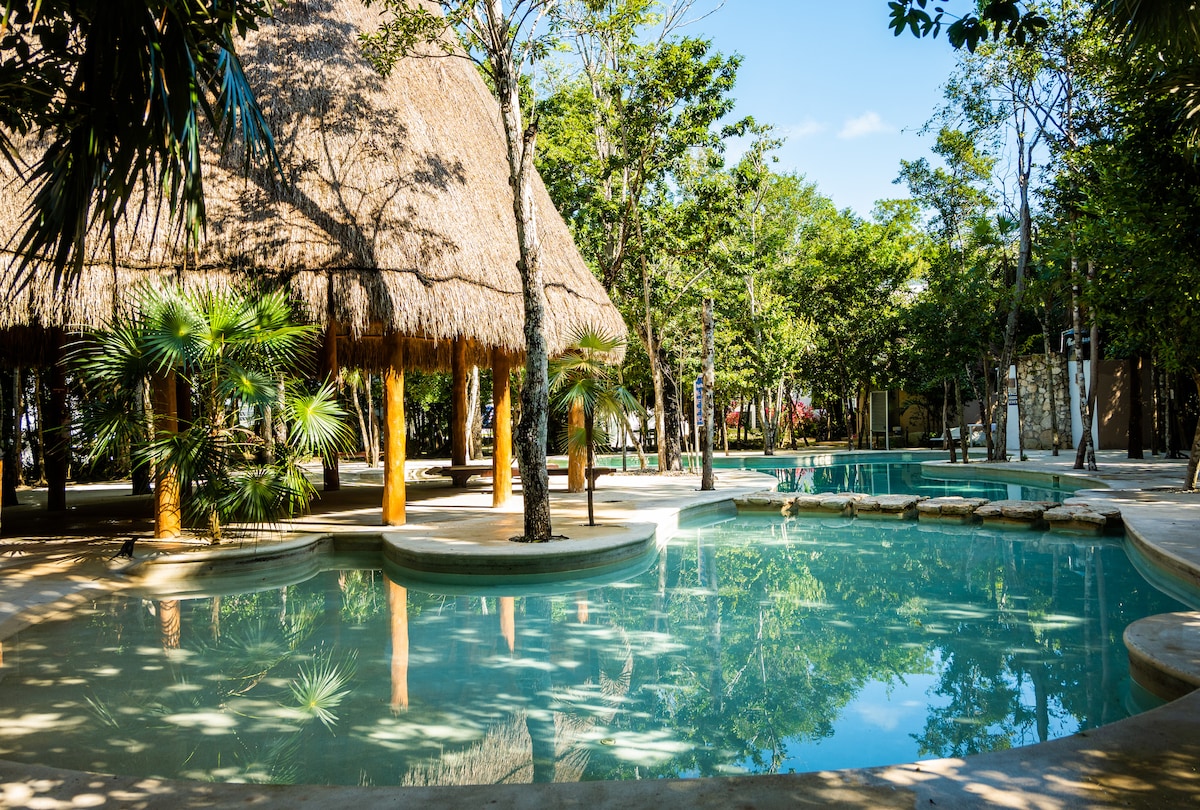 A serene pool area is framed by lush greenery and tropical trees. A thatched-roof structure provides shade, while calm water reflects the surrounding vegetation. Pathways lead around the pool, inviting relaxation in a tranquil setting.