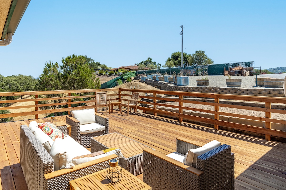 A spacious deck features a comfortable seating arrangement with wicker furniture and a wooden table. Surrounding views include trees and distant structures, creating a serene outdoor setting for relaxation. The clear sky adds to the tranquil atmosphere.