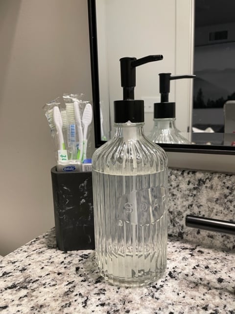 A glass soap dispenser filled with liquid soap is placed on a granite bathroom countertop. A container holding toothbrushes is also visible, with colorful toothbrushes slightly protruding. A mirror is positioned above the counter, reflecting the scene.