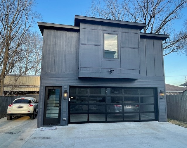 A modern two-story structure features a sleek black exterior with large glass garage doors. A single window is positioned above the garage, allowing natural light to enhance the facade. Nearby, cars are parked in the driveway, surrounded by trees that create a tranquil atmosphere.