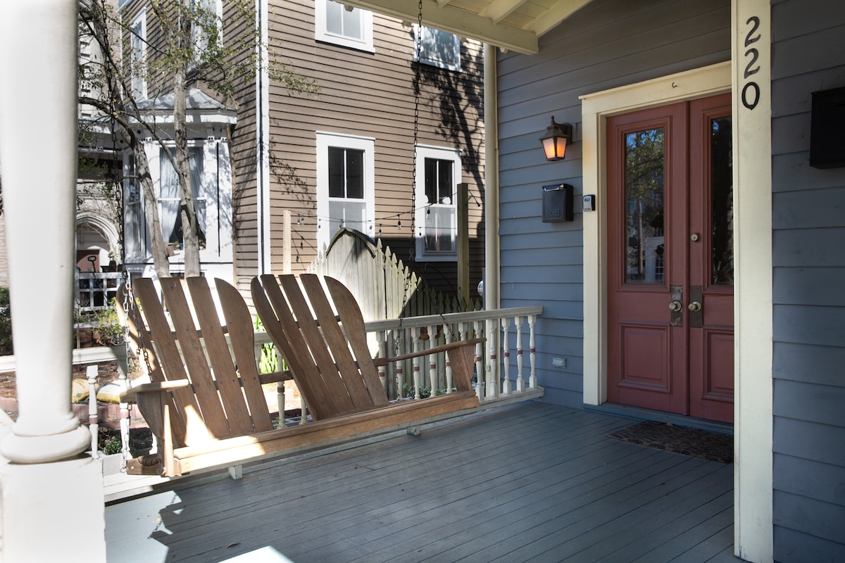 A welcoming front porch features a long, slatted bench, inviting relaxation. The entrance is highlighted by a pair of decorative doors, with a warm light fixture mounted beside them. Additional details include surrounding greenery and charming architectural elements of the historic home.