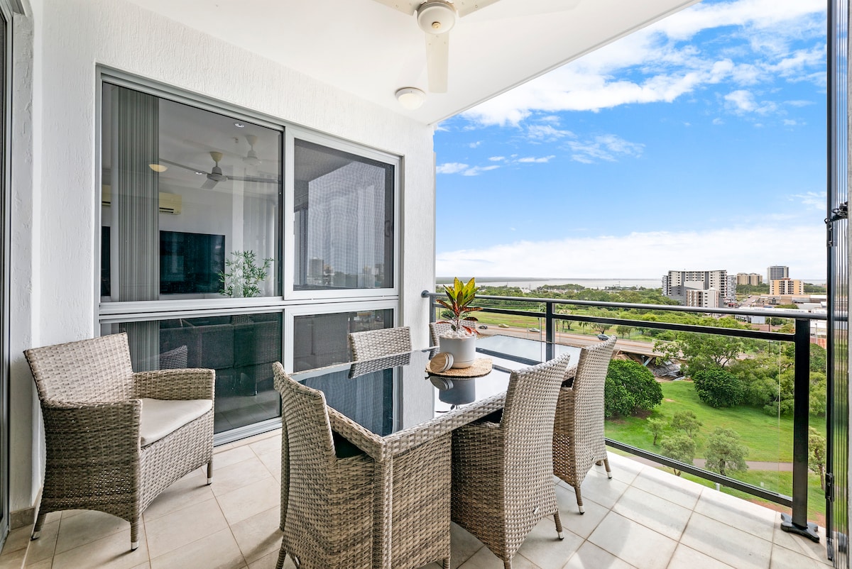 A covered balcony features a large outdoor dining table surrounded by wicker chairs. Greenery is visible in the surrounding landscape, with expansive city views in the distance. Natural light brightens the space, providing an inviting area for alfresco meals.