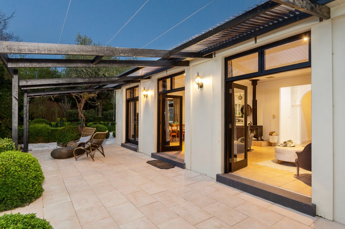 An outdoor patio area features large double doors leading into the interior spaces. Cozy seating arrangements are positioned beside manicured gardens, with soft lighting illuminating the entrance. Greenery surrounds the space, creating an inviting transition from indoors to the serene outdoor surroundings.