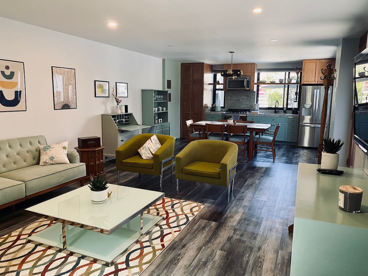 A modern living area is presented with a light green sofa and two olive green chairs, accompanied by a glass coffee table. A patterned rug adds color, while a dining area with a wooden table and chairs is visible in the background. Natural light flows through large windows.