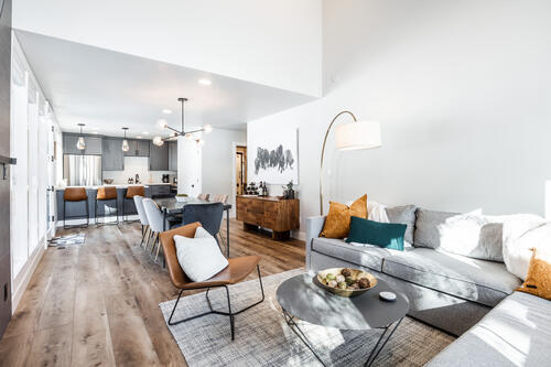 An open living area features a light-colored sofa adorned with decorative pillows and a round coffee table. A dining area with modern bar stools is visible, while a wooden sideboard displays tasteful decor. Natural light brightens the space through large windows.