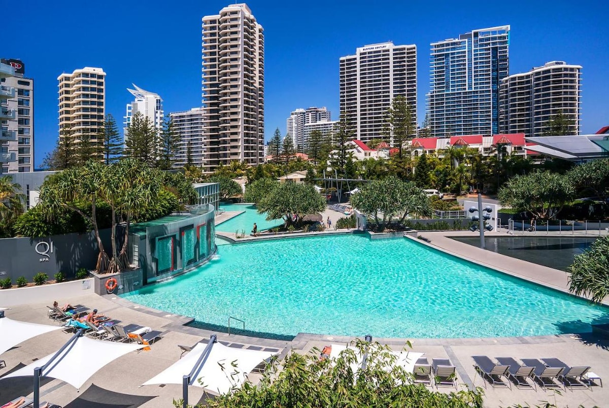 A large outdoor pool is surrounded by manicured landscaping and sun loungers. Towering high-rise buildings provide a backdrop against a clear blue sky. Lush greenery adds to the serene atmosphere, inviting relaxation and enjoyment of the refreshing water.