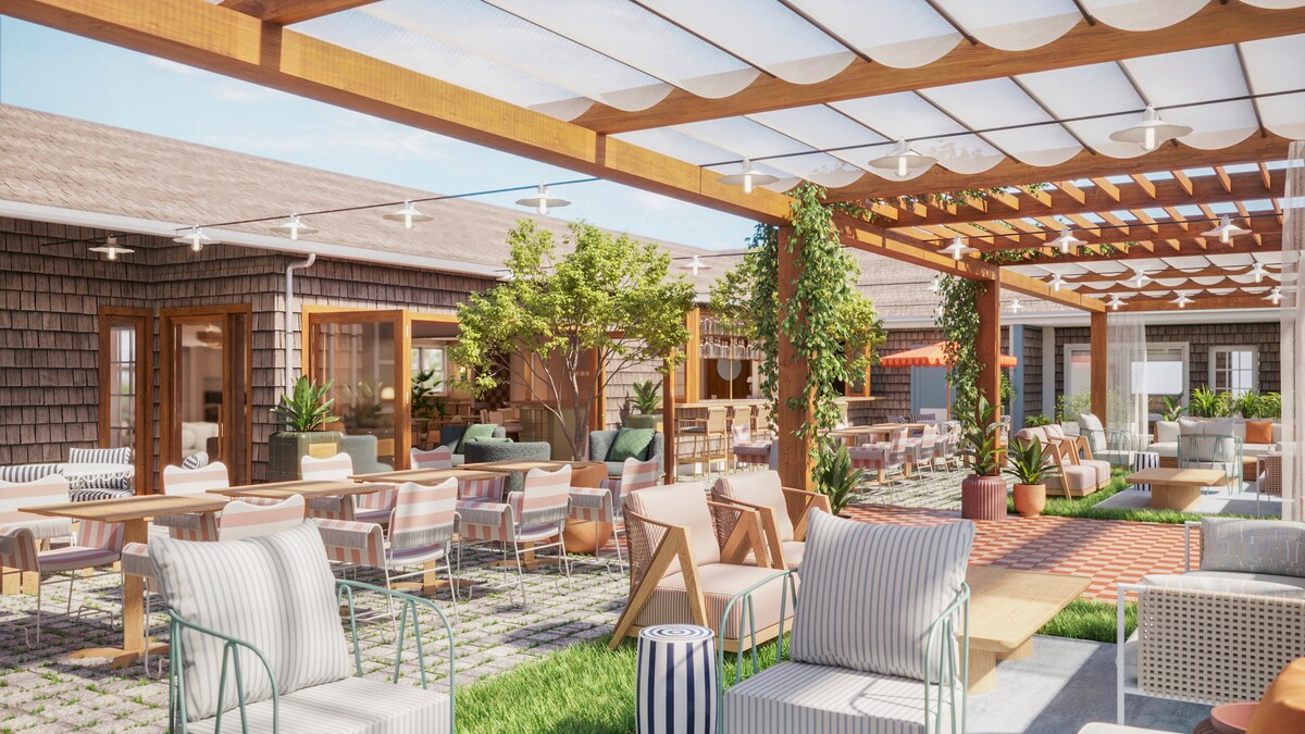 An outdoor dining area is surrounded by lush greenery and wooden structures. Comfortable seating options, including lounge chairs and tables, are arranged under a canopy. The space is brightened by natural light, creating an inviting environment for relaxation and social gatherings.