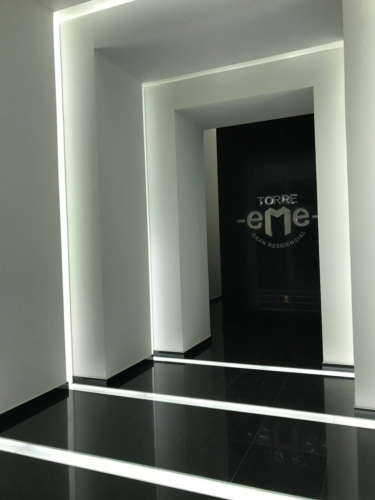 A modern hallway is highlighted by sleek, minimalist design. Bright white light accents the clean lines of the architectural features, while a dark entrance at the end leads to the residential area. The polished black floor reflects the light, enhancing the space's contemporary feel.