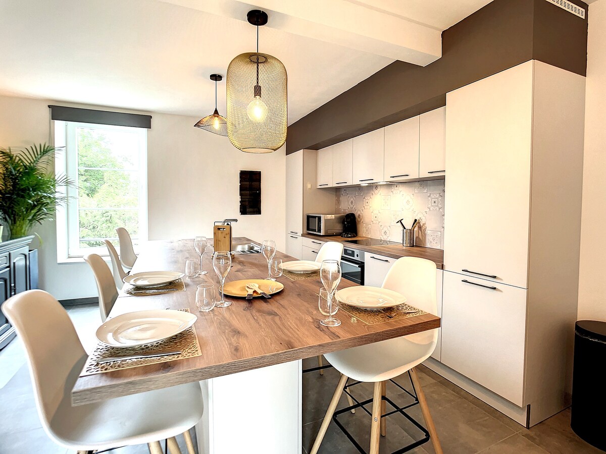 The kitchen area features a long wooden table set for dining, with plates and glasses positioned neatly. Modern appliances are visible, including a microwave and a coffee maker. Large windows provide natural light, illuminated by pendant lighting above the table.