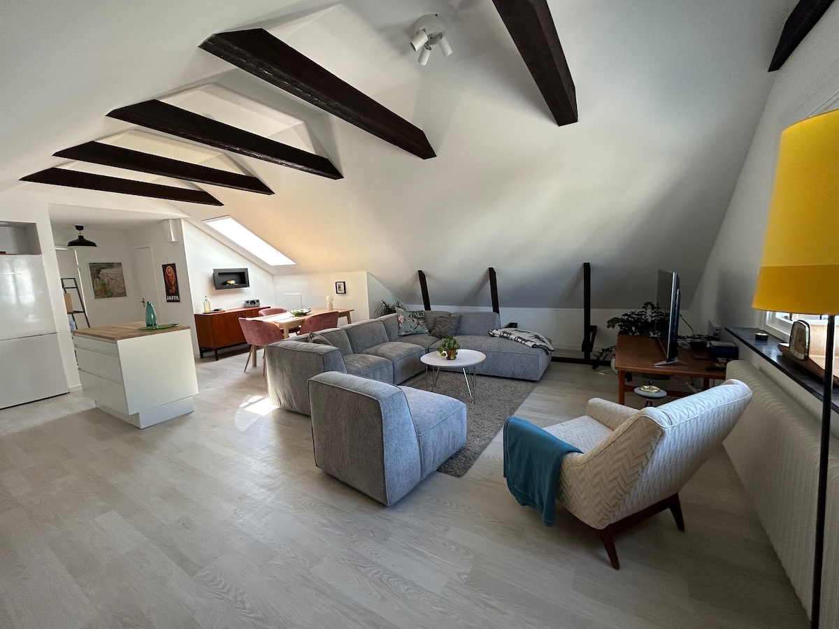 A spacious living area features wooden beams overhead and large windows providing natural light. A sectional sofa and a single armchair are arranged around a central coffee table on a soft area rug. A television is positioned against the wall, enhancing the comfort of the space.