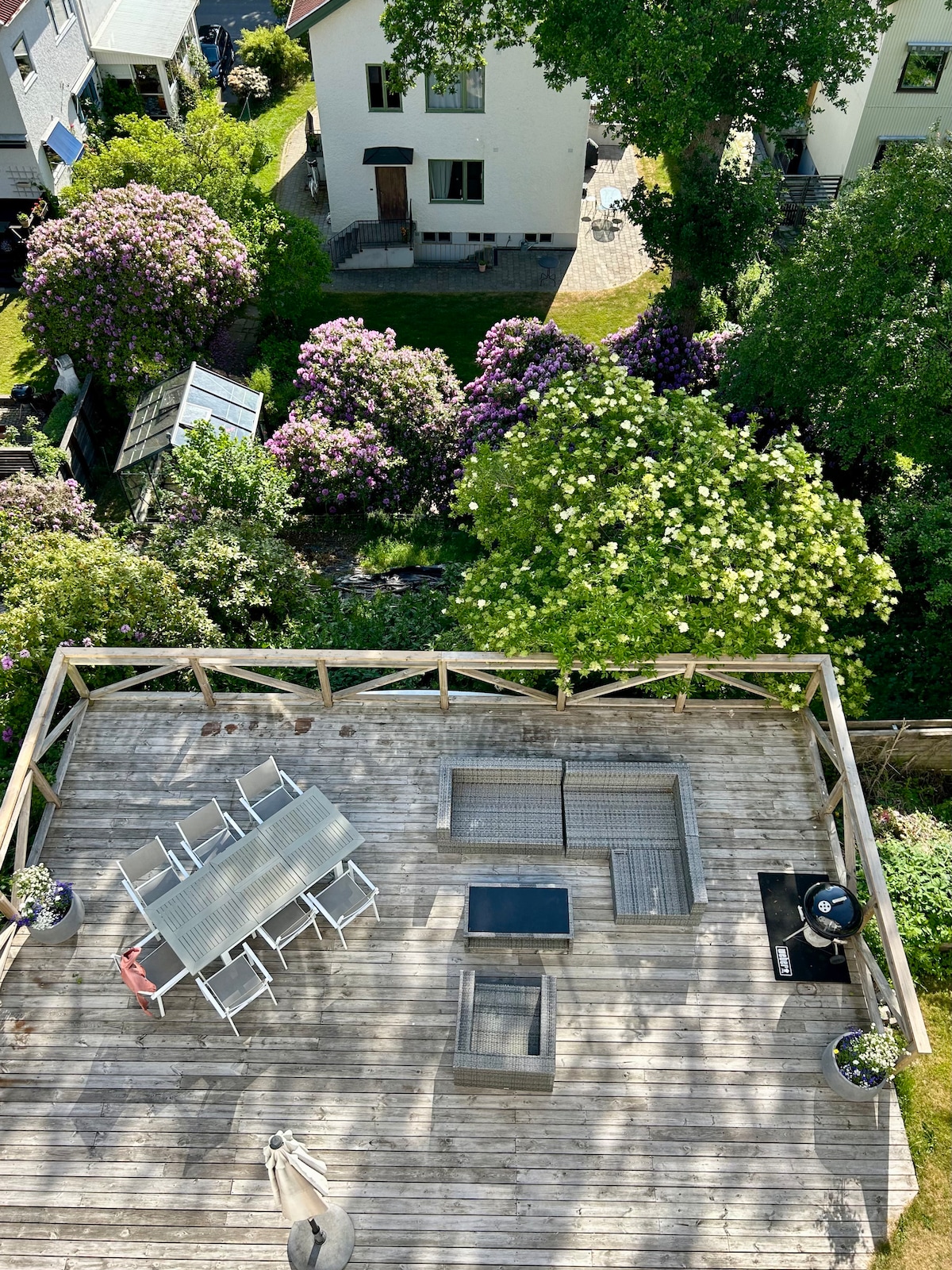 A spacious wooden terrace offers an outdoor seating area with metal furniture and a table surrounded by potted plants. Below, a lush garden features blooming flowers and green trees, providing a serene landscape view.