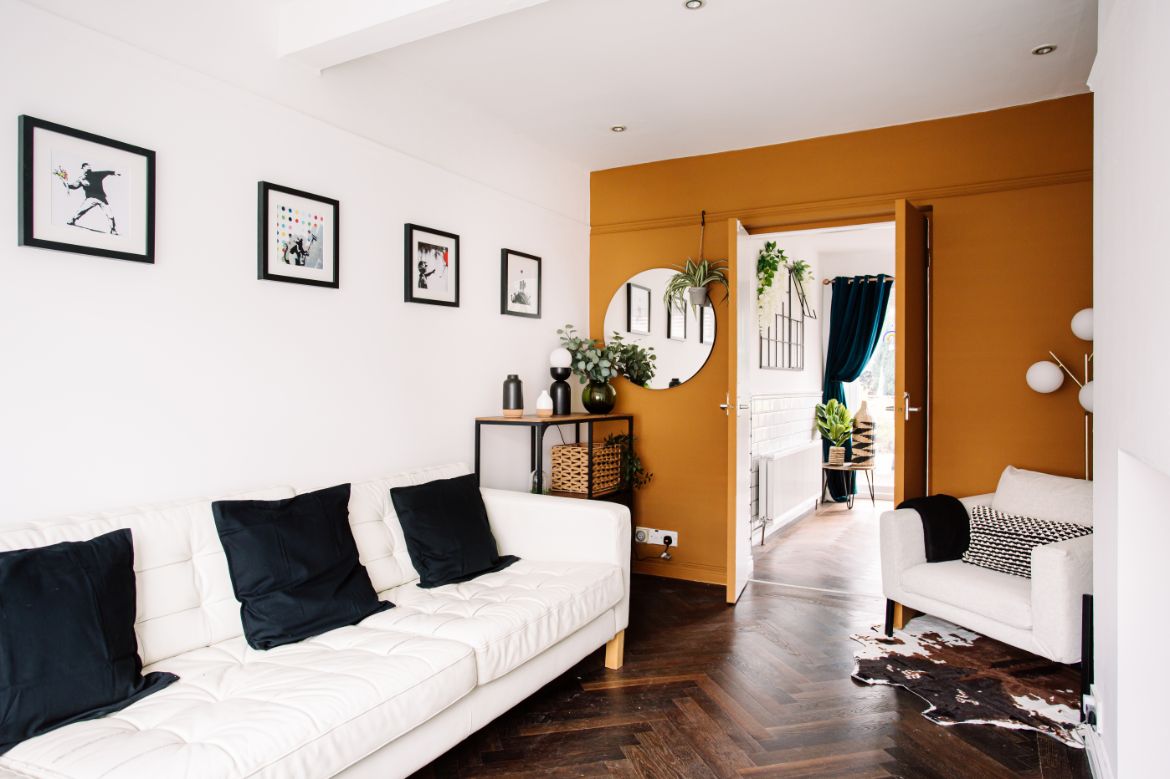 A bright living area features a white leather sofa accented with black cushions, complemented by framed artwork on the walls. A cozy armchair and decorative elements add character, while the space transitions to a hallway adorned with plants and contrasting wall colors.