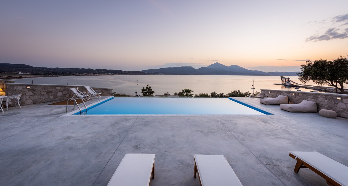 A serene swimming pool reflects the evening sky, surrounded by a smooth stone deck. Sun loungers are positioned nearby, offering a space for relaxation. The view includes a distant mountain range and coastline, creating a tranquil backdrop for sunset moments.