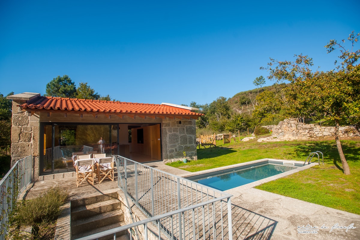 A stone house with a red-tiled roof is set amid a natural landscape. An outdoor pool is visible in the foreground, surrounded by grass, with lounge chairs nearby. Large windows offer views into the living area, enhancing the connection with the outdoors.
