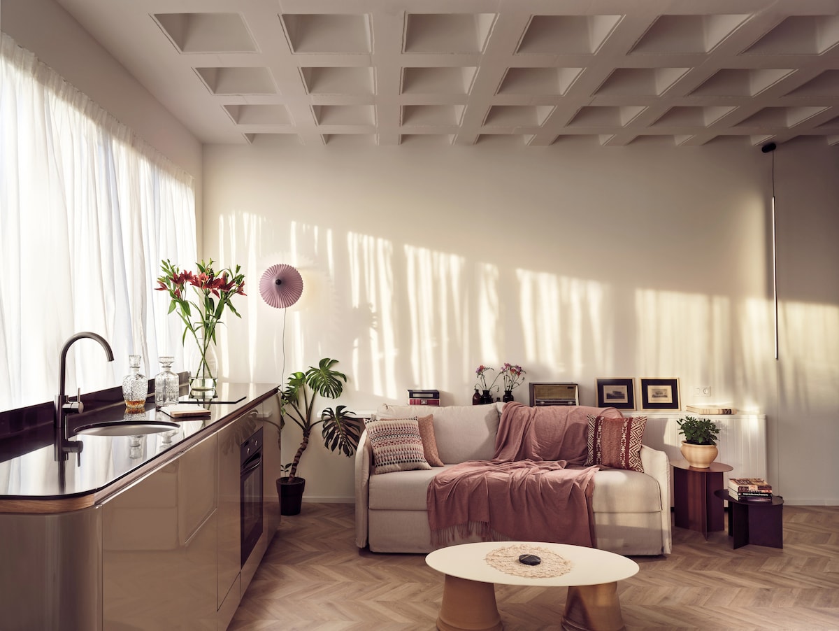 A light-filled living space features a sofa adorned with a cozy blanket and decorative cushions. A coffee table sits in the center, complemented by potted plants. A modern kitchen area is visible, with sleek cabinetry and natural light filtering through sheer curtains.
