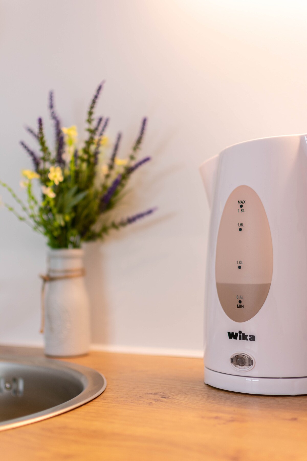 A kettle is positioned on a wooden countertop, featuring clear measurement markings. Nearby, a small vase holds vibrant flowers, adding a touch of color to the kitchen space. The setup emphasizes functionality and simplicity in the setting.