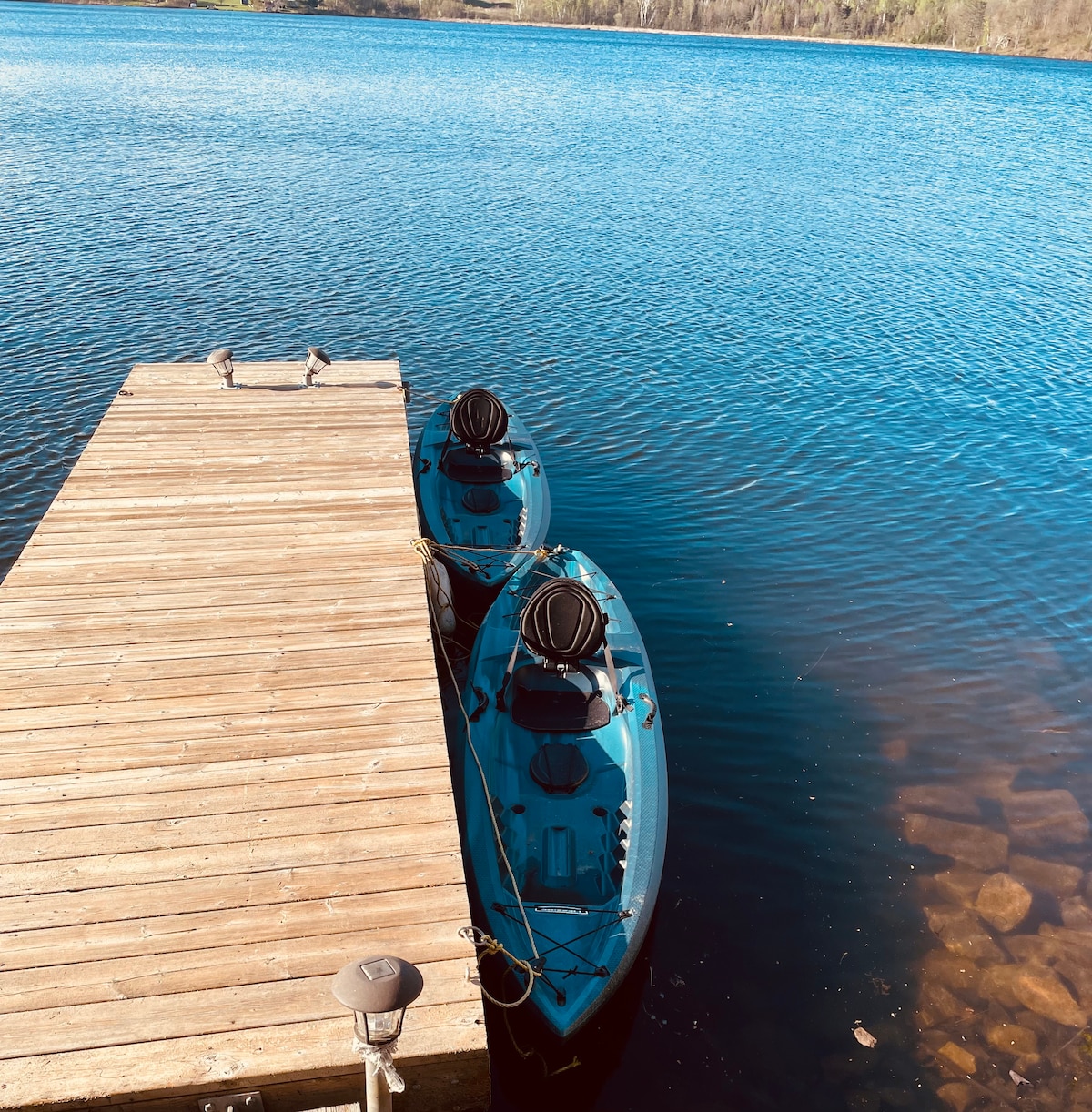 A wooden dock extends over the calm waters, featuring two kayaks securely tied alongside. The bright blue hulls of the kayaks contrast with the tranquil shades of the water, surrounded by a natural landscape in the distance.