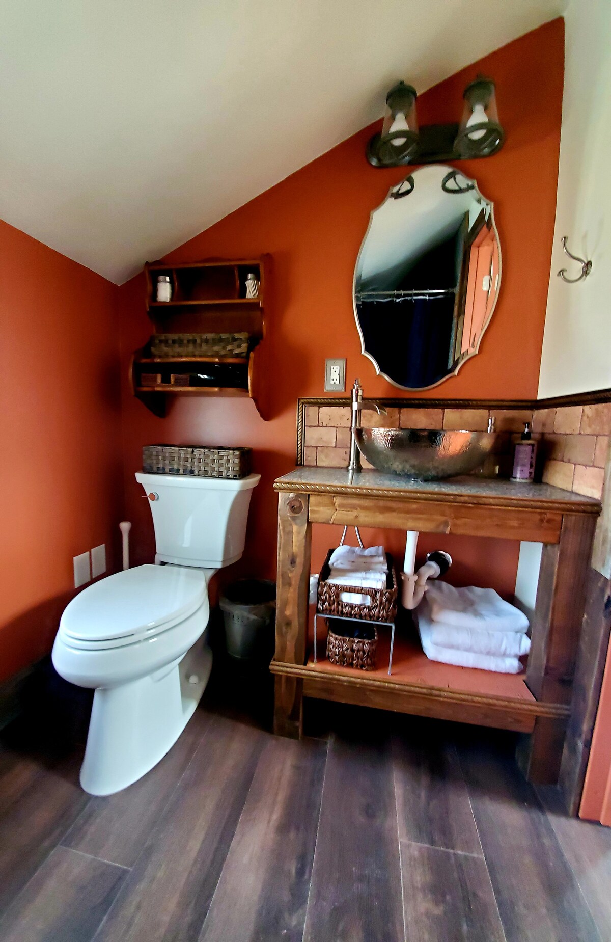 A compact bathroom features rich orange walls complemented by natural wood accents. A modern toilet is positioned beside a wooden vanity with an oval sink. Above the sink, a decorative mirror is supported by wall sconces. Towels and toiletries are neatly arranged on the shelves and vanity.