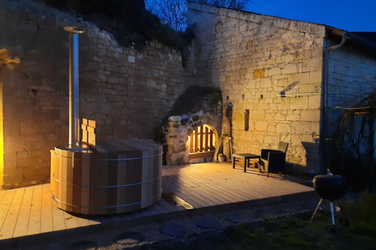 An outdoor terrace features a wooden Nordic bath, quietly illuminated by warm lighting. A simple chair sits nearby, while part of a stone wall adds rustic charm to the serene setting. The wooden deck enhances the inviting atmosphere of the space.