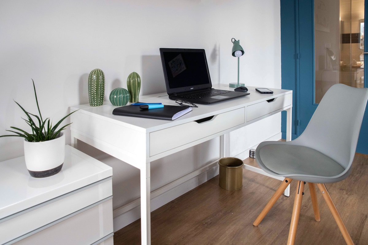 A workspace is provided featuring a clean white desk adorned with small potted plants and a laptop. A sleek grey chair complements the modern design. A green lamp illuminates the area, with a notebook and pen positioned neatly on the desk.