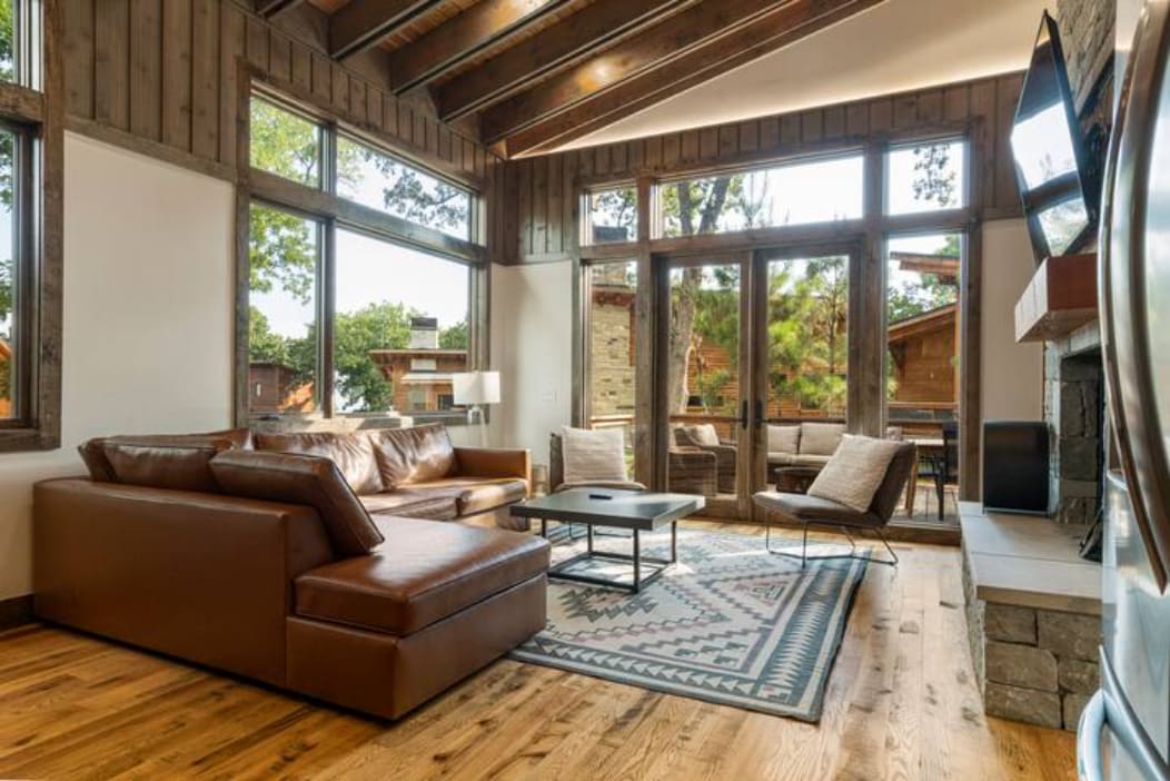 A spacious living area features a large sectional sofa with rich brown leather. A coffee table sits on a patterned rug, while floor-to-ceiling windows provide abundant natural light and views of the surrounding trees. A stone fireplace adds a rustic touch to the modern decor.