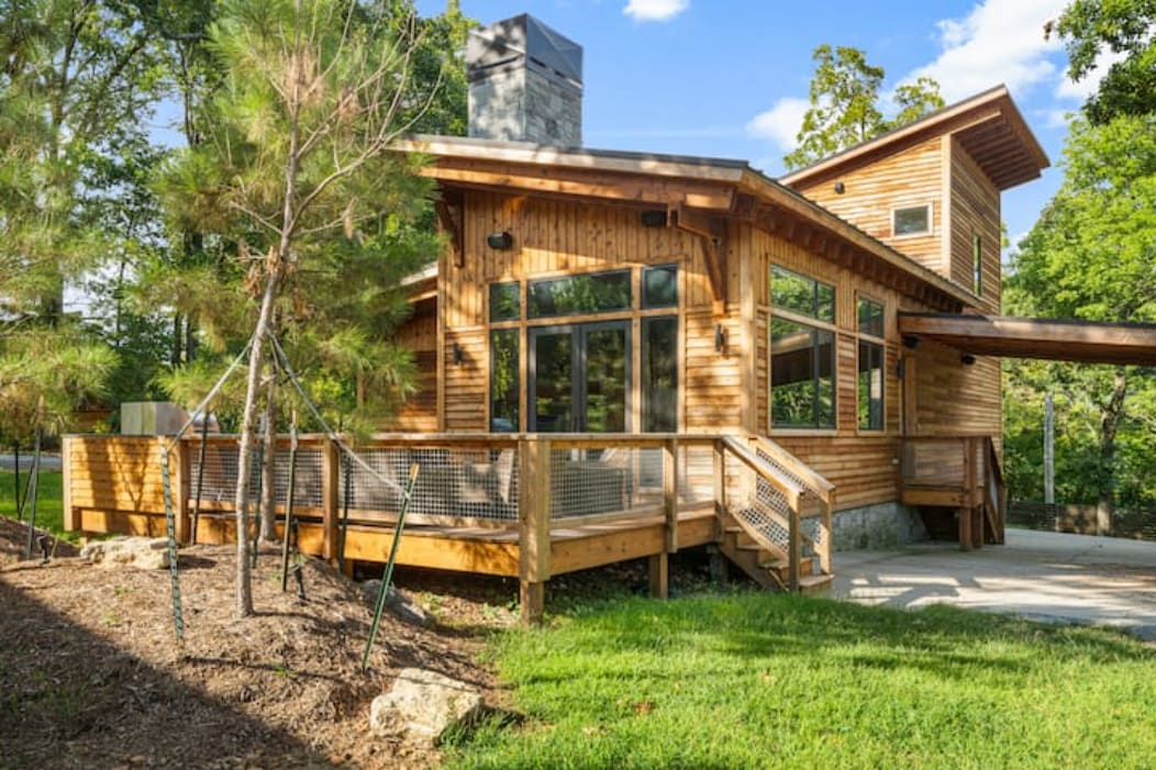A modern rustic cabin is presented with a wooden exterior and large, expansive windows. A spacious wrap-around deck enhances outdoor access, while the surrounding greenery provides a natural setting. The structure showcases a blend of contemporary design and rustic charm.