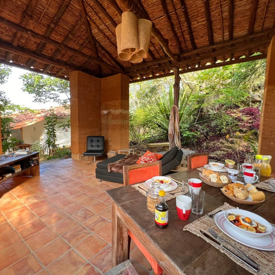 A spacious outdoor seating area features a dark sofa and wooden dining table set, arranged with breakfast items including plates, glasses, and a bottle. Lush greenery is visible in the background, complementing the natural tones of the structure's materials.