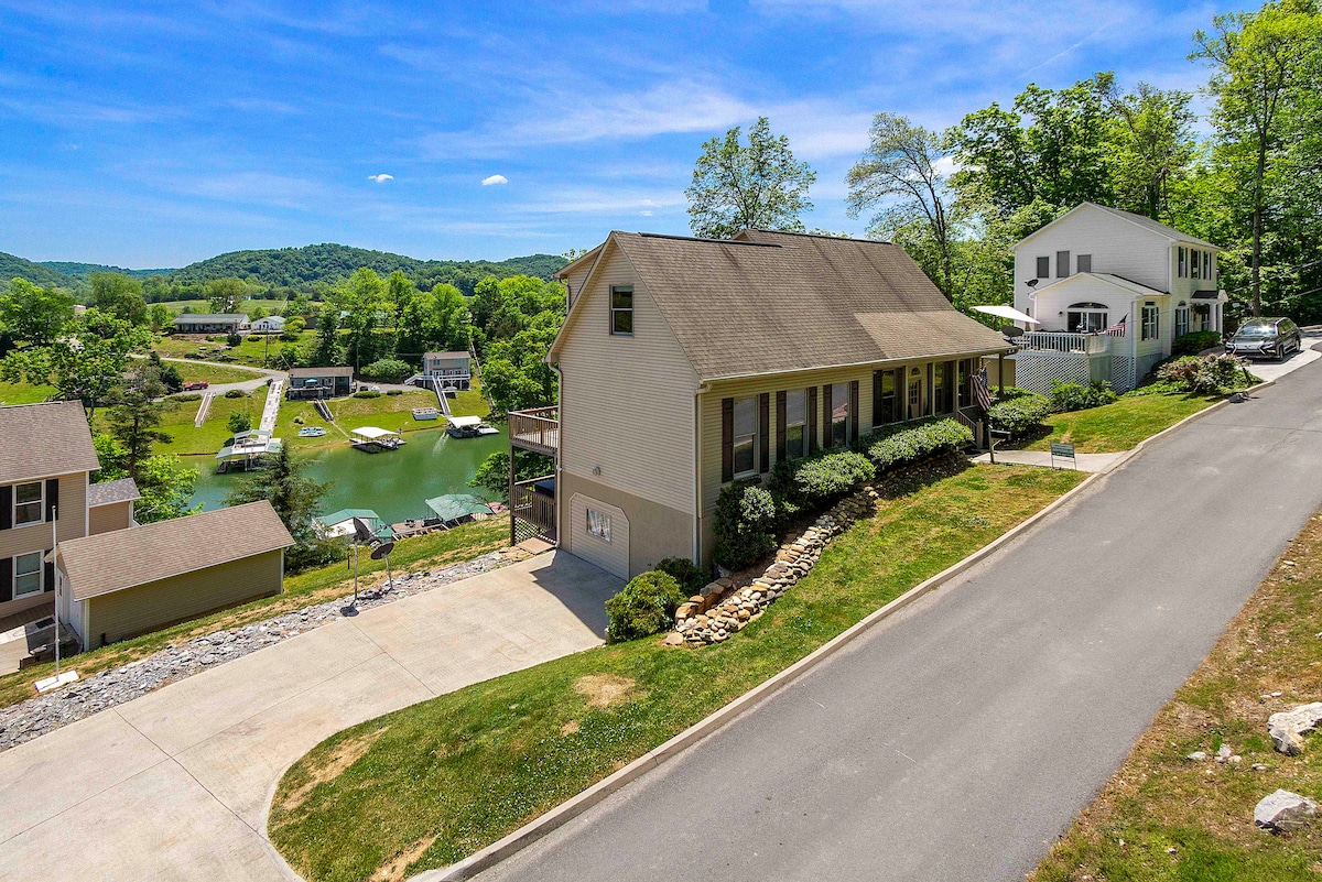 The image captures a welcoming lakefront home nestled among lush green trees. The house features a light exterior and is positioned on a gently sloping driveway. A view of the serene Norris Lake and adjacent dock area is visible from this elevated perspective.