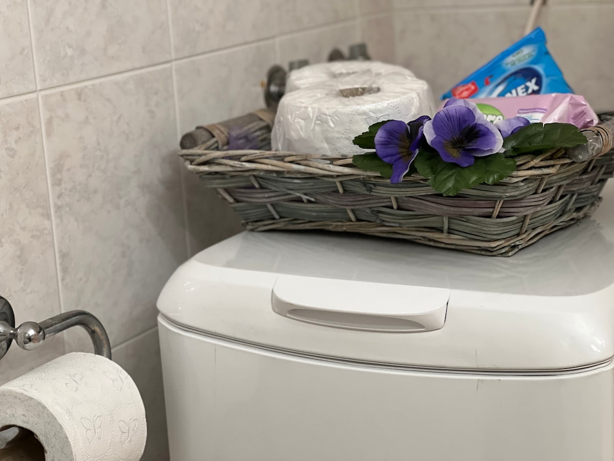 A bathroom corner features a neatly arranged basket atop a washing machine. The basket contains toiletries, including paper rolls and cleaning supplies, alongside decorative flowers. A roll of toilet paper is positioned on a nearby holder, enhancing functionality.