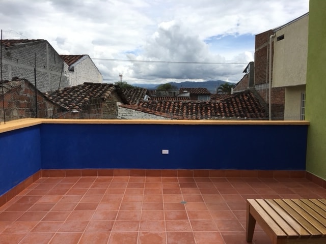 A spacious terrace is showcased, featuring a tiled floor and a wooden bench. The vibrant blue wall contrasts with the earthy tones of the surrounding rooftops. Clouds are visible in the sky, enhancing the open ambiance of the space.