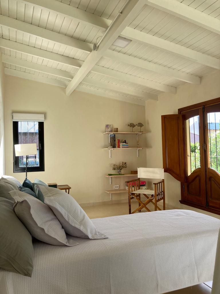 A bright bedroom featuring a spacious bed with layered bedding and decorative pillows. A small reading nook includes a chair and side table, while shelves display decorative items. Large windows invite natural light and offer views of the surrounding greenery.