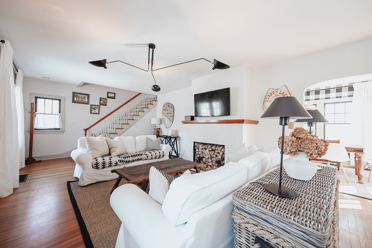 An inviting living room features a large, comfortable sectional sofa arranged around a wooden coffee table. A modern light fixture hangs overhead, and a television is mounted above a white brick fireplace. Natural light streams through large windows adorned with sheer curtains.