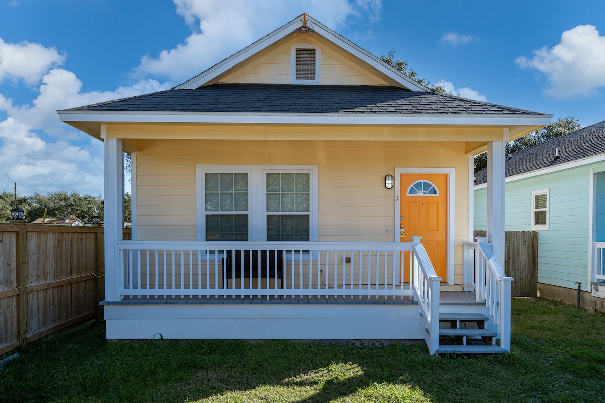 A cottage exterior is presented with a welcoming orange front door and white railings along the porch. Two large windows are framed on either side of the door, and a well-maintained lawn surrounds the home. The sky is clear with scattered clouds.