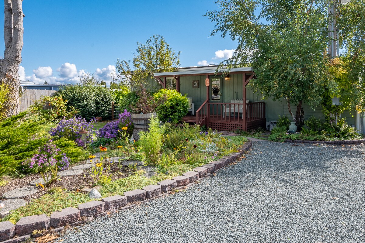 A charming green exterior of a bungalow is framed by vibrant landscaping, featuring various shrubs and flowering plants. A gravel pathway leads to the entrance, while trees and a low fence add to the inviting setting under a clear blue sky.