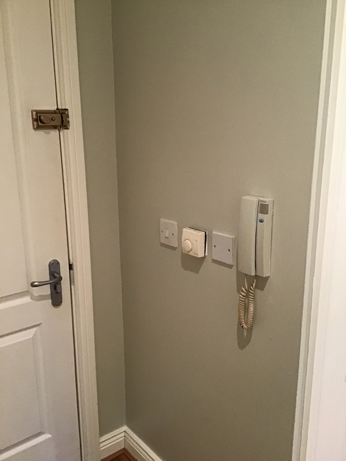 The image shows a corner of a room featuring a light-colored wall. A door is visible on the left side, equipped with a lock. On the wall, a thermostat is installed alongside a phone intercom, indicating connectivity and comfort.