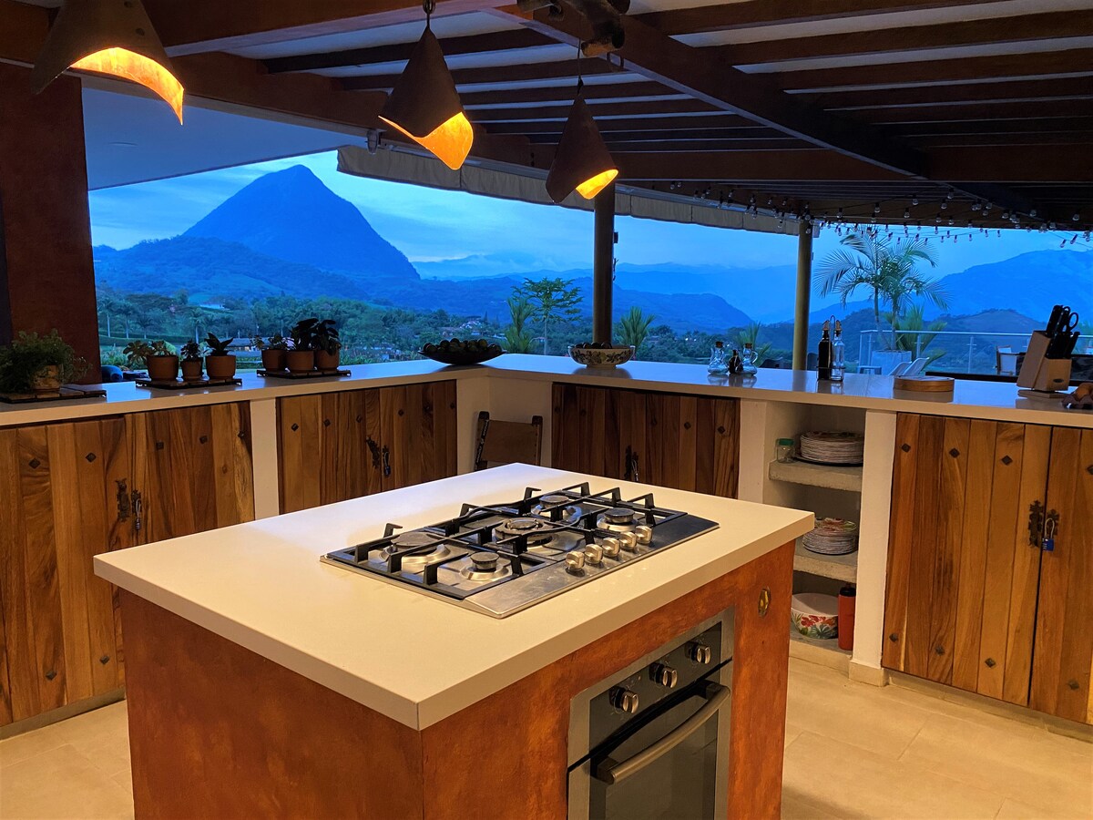 An open kitchen features a central island with a gas cooktop and an oven beneath. Wooden cabinetry lines the space, complemented by pendant lighting above. Through large windows, panoramic views of the surrounding mountains and greenery are visible, creating a spacious and airy environment.
