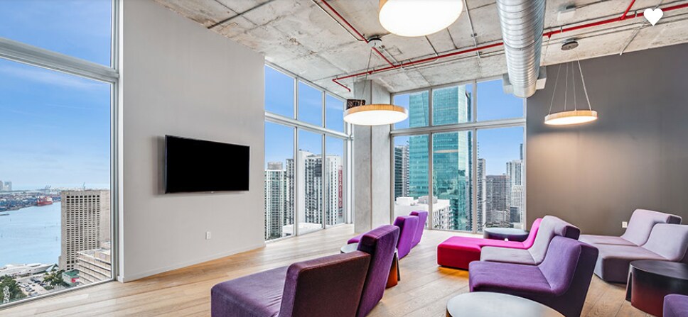 A bright lounge area features large windows that provide expansive views of the city and water. Colorful seats in purple and pink are positioned around a simple wooden table, and a flat-screen TV is mounted on the wall, enhancing the inviting atmosphere.