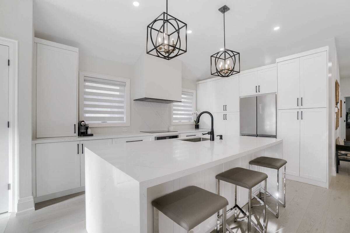 The kitchen features a large island with a waterfall countertop and seating for three. Modern light fixtures hang above, illuminating the area, while sleek cabinetry and a stainless steel refrigerator complete the contemporary design. Natural light filters through the window treatments, creating a bright space.
