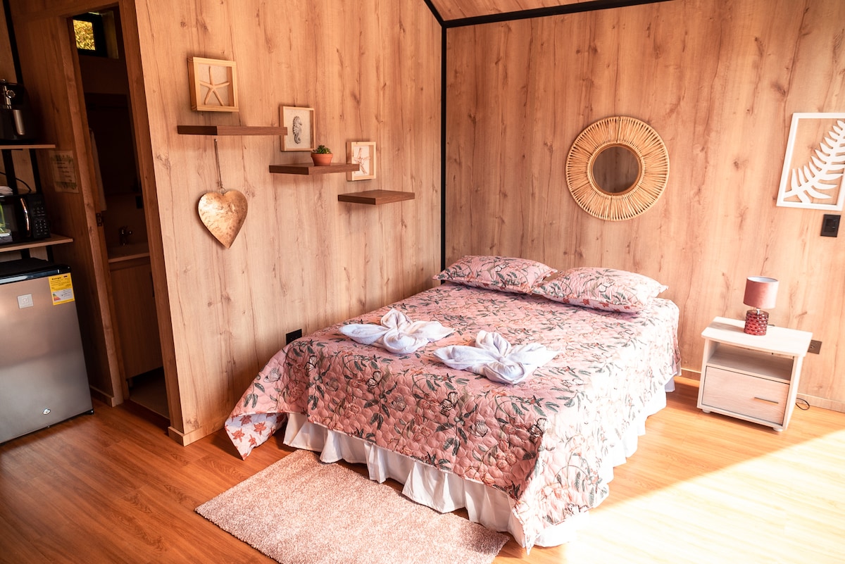 A comfortable sleeping area features a floral-patterned bedspread covering a spacious bed. Soft natural light enters through a window, illuminating wooden walls. A small bedside table and a round mirror enhance the cozy atmosphere, while minimalist decor elements provide a soothing touch.
