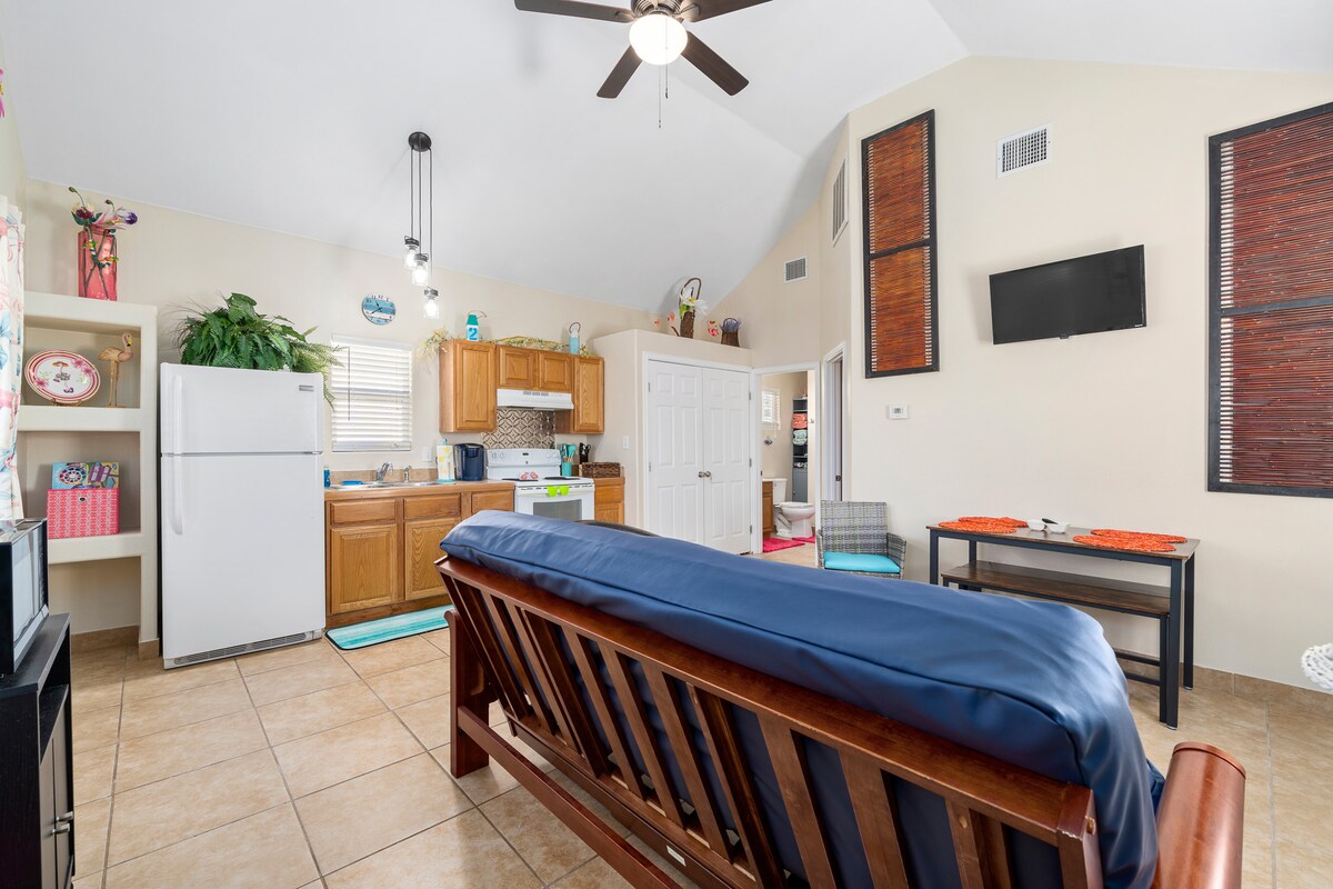 A cozy living space features a wooden futon facing a small dining table and a kitchenette. Bright natural light flows through the windows, and a ceiling fan provides ventilation. Decorative elements include colorful accents and plants, contributing to a lively atmosphere.