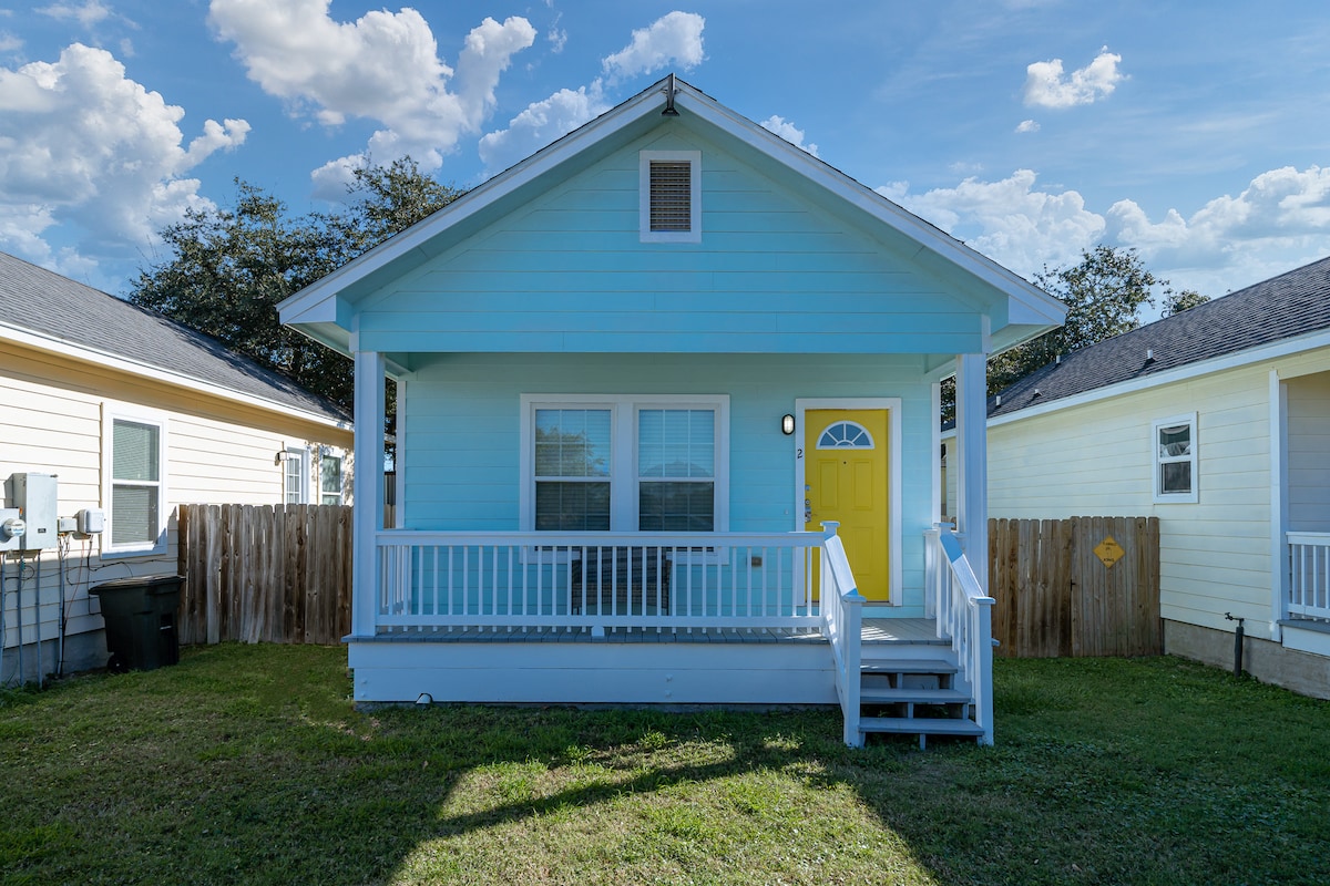 A quaint cottage with a light blue exterior features a cheerful yellow door. The front porch provides steps up to the entrance, surrounded by a grassy yard and wooden fencing. Bright skies and fluffy clouds create a serene backdrop.