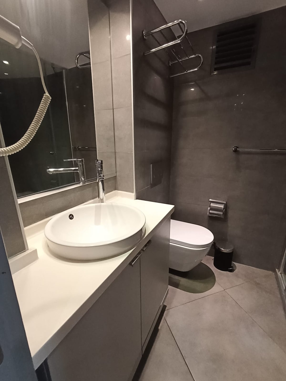 A contemporary bathroom features gray tiled walls and floors. A round sink is positioned above a modern cabinet, with a shower area visible through glass doors. A toilet is placed discreetly beside the shower, and towel racks are mounted on the wall.