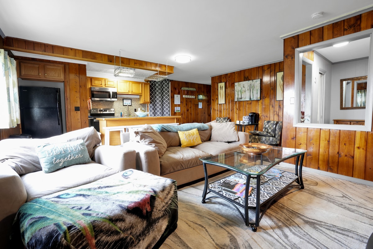 A spacious living room features a comfortable oversized couch and a recliner, arranged around a glass coffee table. Natural wood paneling decorates the walls, complemented by a well-equipped kitchen visible in the background. Light enters through nearby windows, enhancing the overall ambiance.