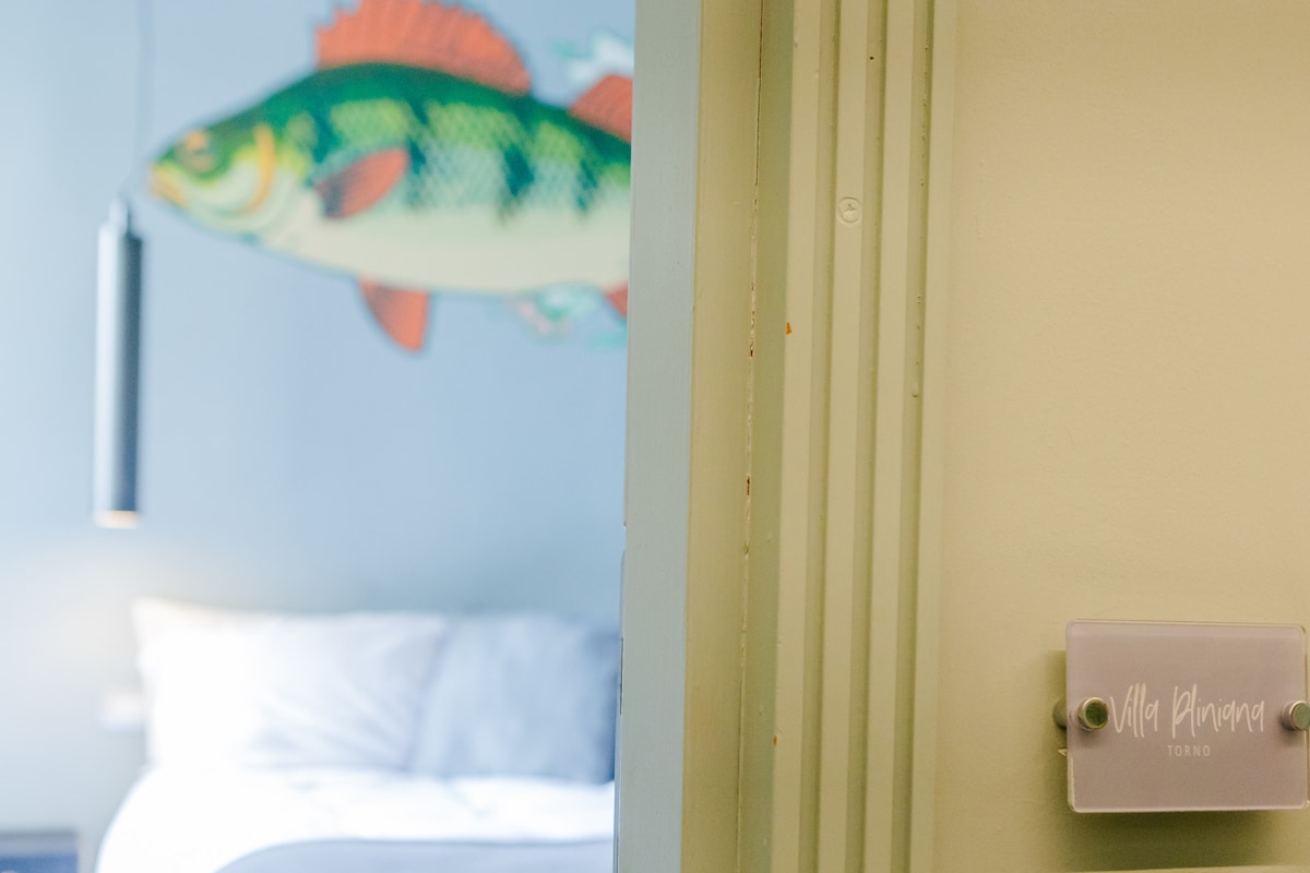 A colorful wall mural of a fish is visible in the background through an open doorway. The light-colored walls complement the bright decor. A glimpse of a spacious bed can be seen, suggesting a cozy sleeping area beyond.